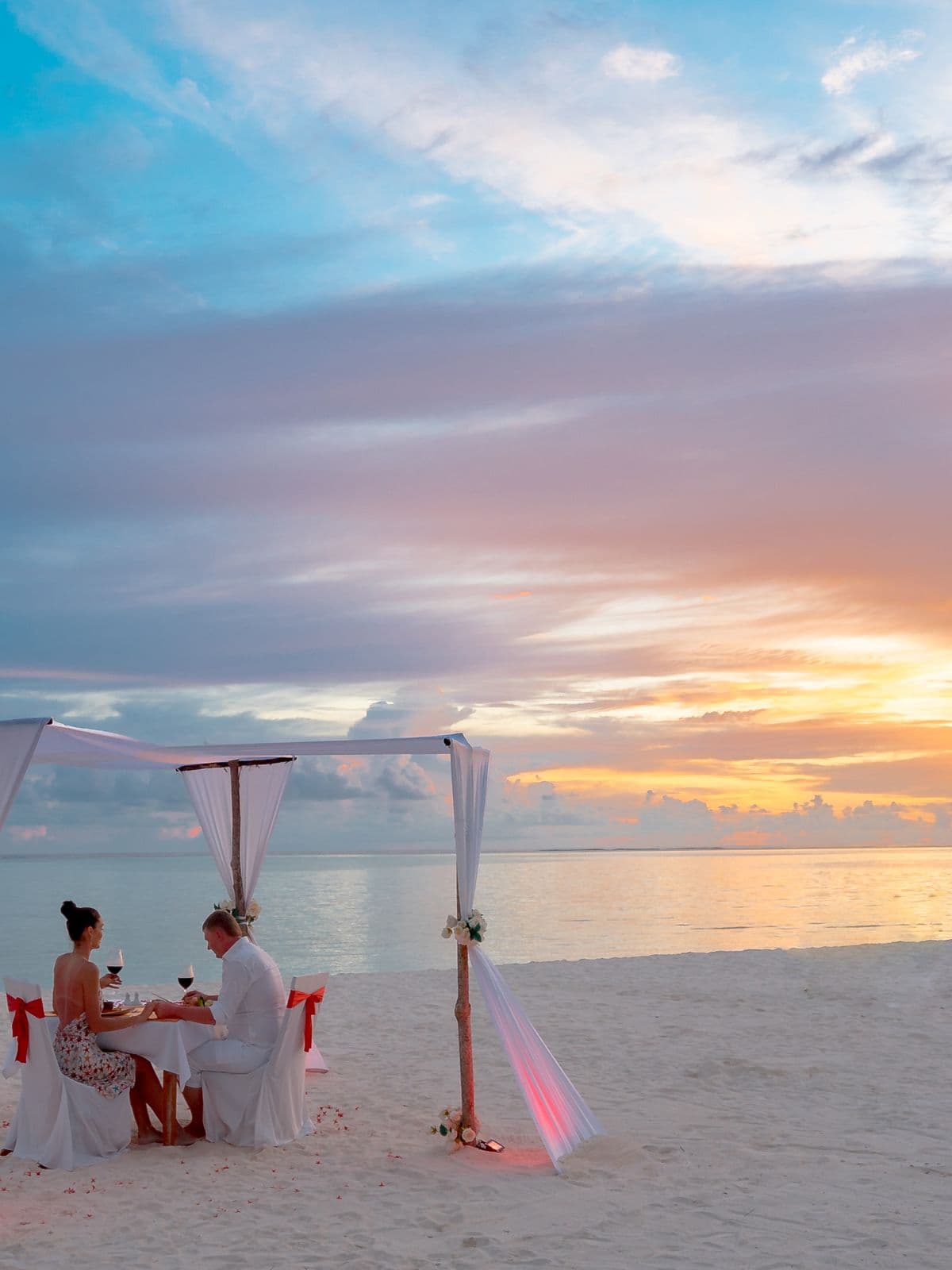 A table laid for dinner under a light canopy on the sand at sunset