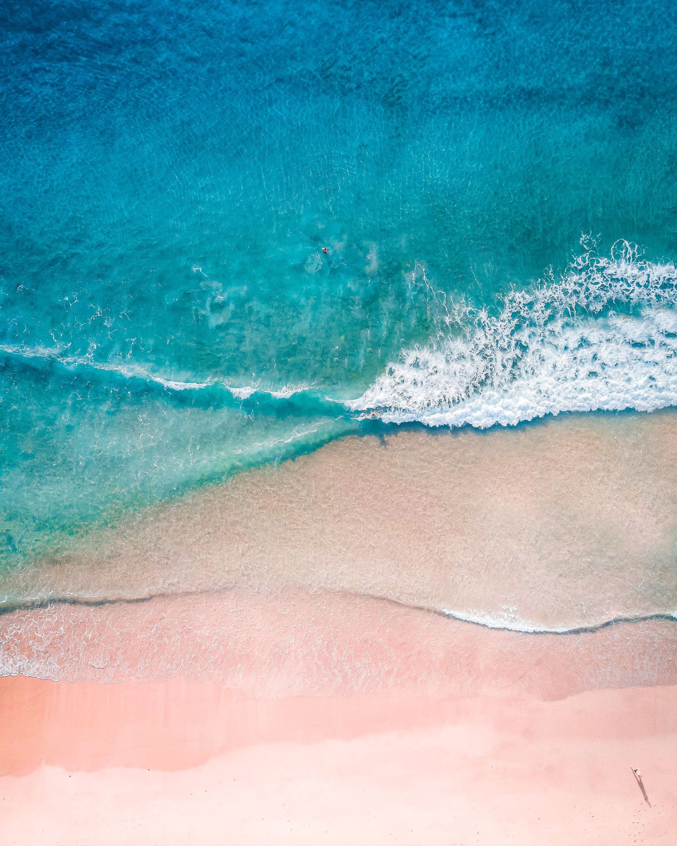 Turquoise water and white surf breaking on a pale, empty beach, seen from above
